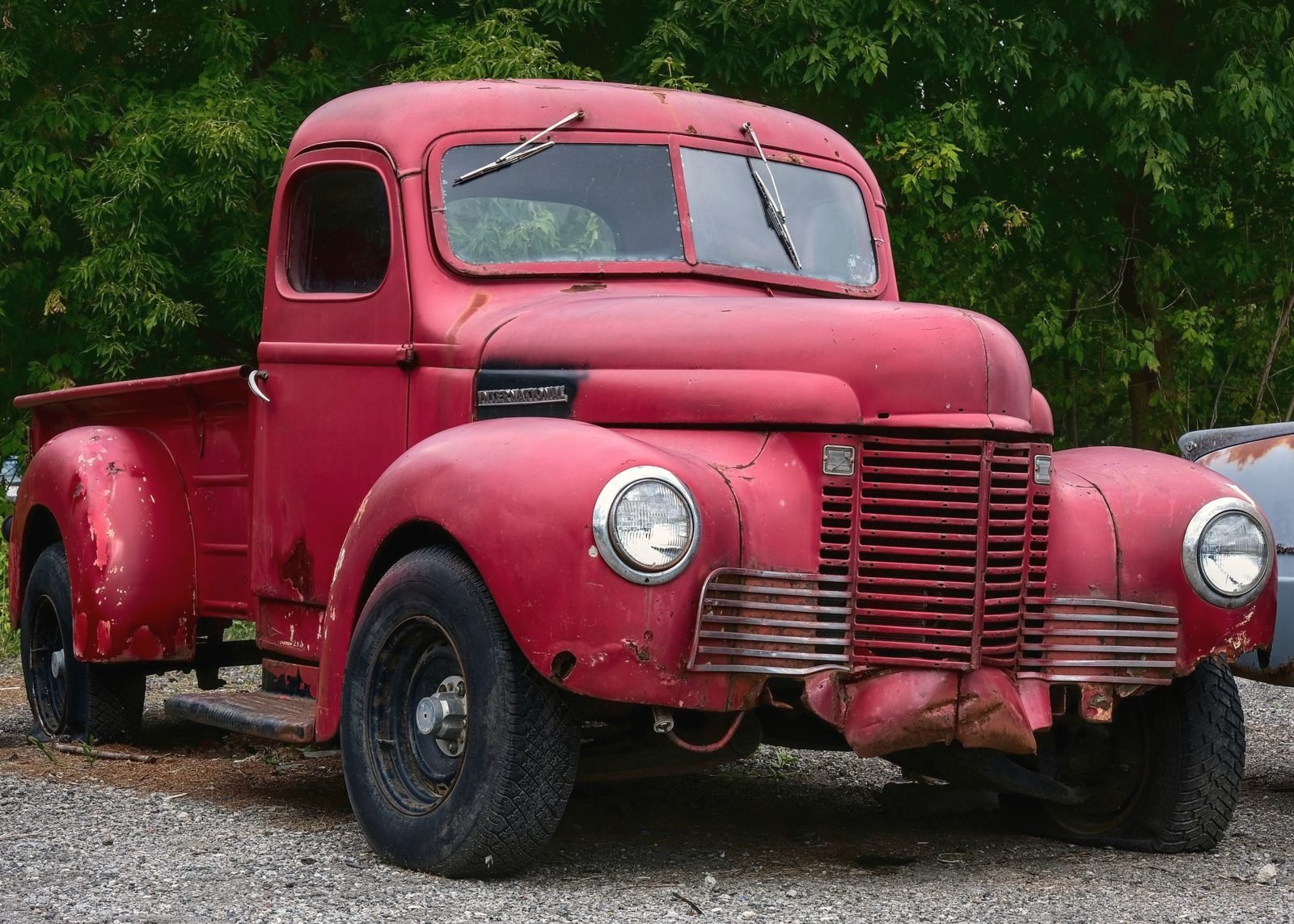 A faded red vintage pickup truck parked on gravel under trees
